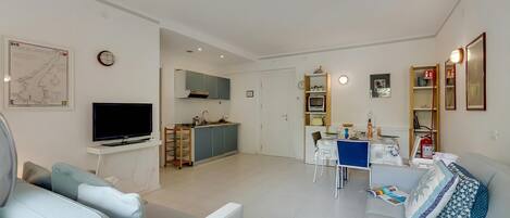 A modern living room with a flat-screen TV, a sofa, a dining table, and a kitchen area in the background.