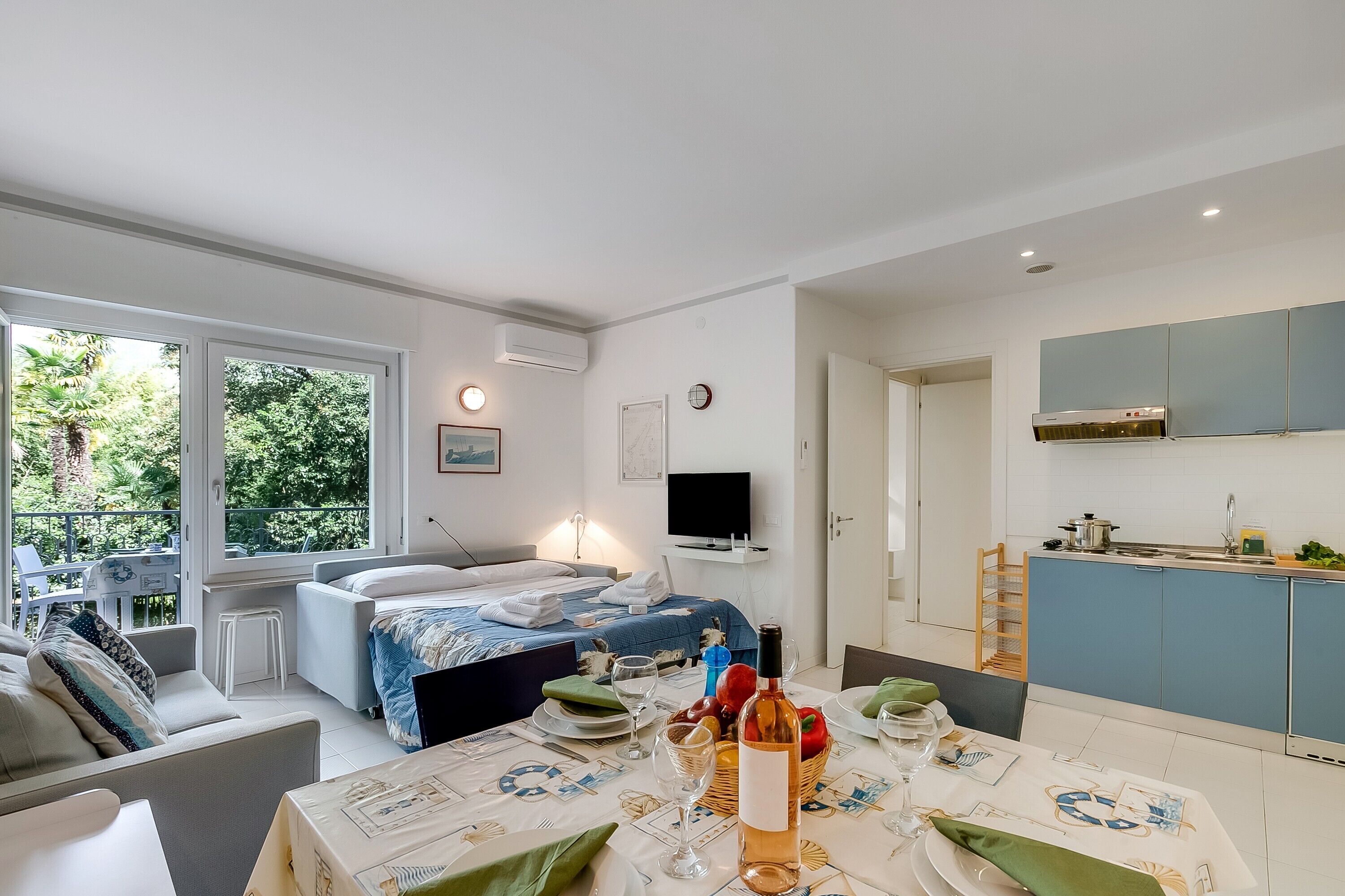 A modern hotel room with a large bed, a dining area with a table set for two, a kitchen area, and a balcony with a view of greenery.