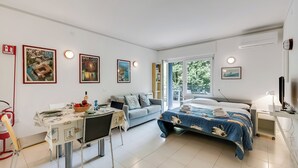 A modern hotel room with a bed, a desk, a sofa, and a dining table. There are framed pictures on the wall and a window with a view of trees. - Riva Lago e Spiaggia Apartment 3 (Riva del Garda)