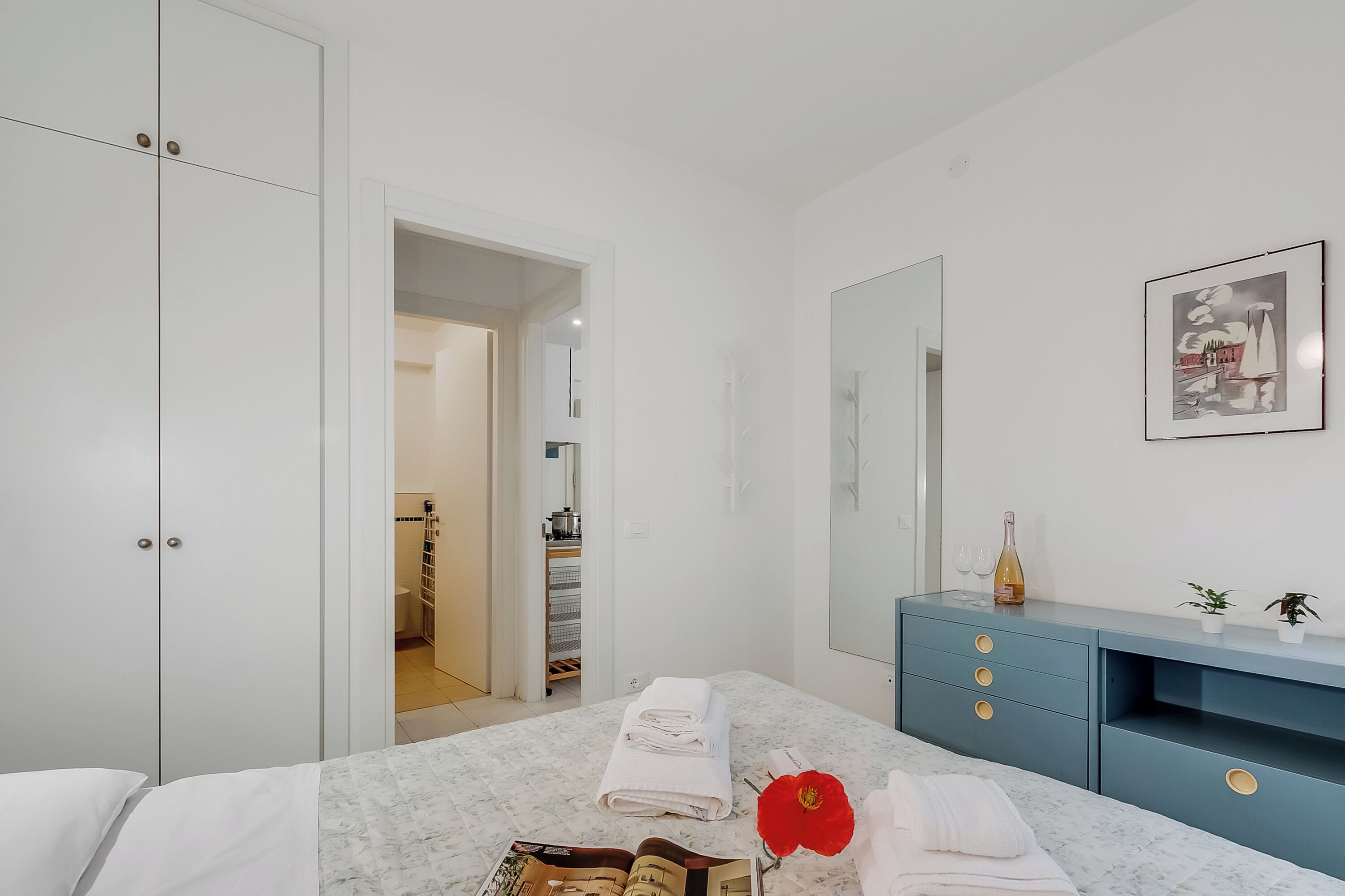 A hotel room with a bed, a vanity with a mirror, a blue dresser, and a framed picture on the wall.