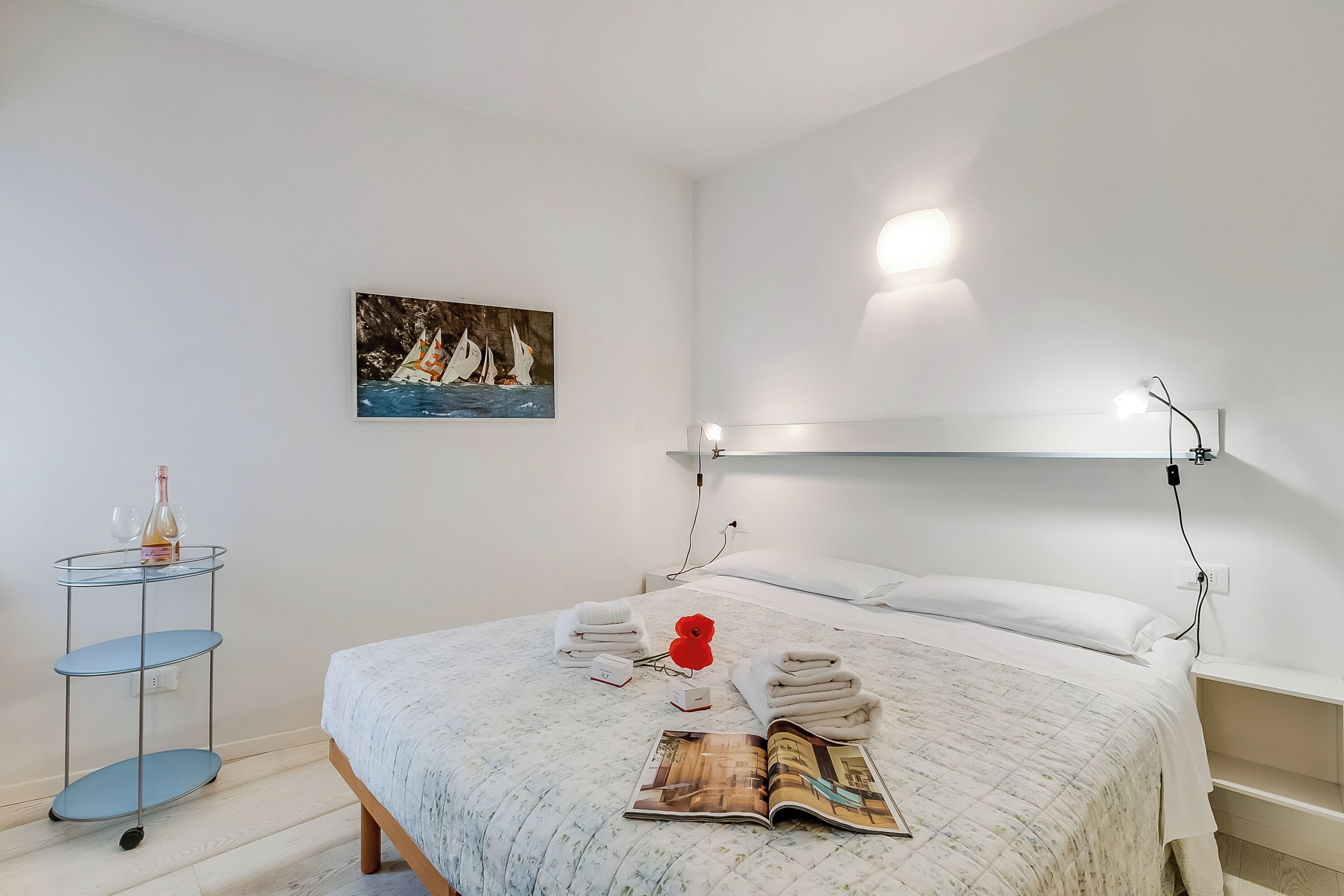 Een opgemaakt bed met een sprei, een tijdschrift en handdoeken tegen een witte muur.