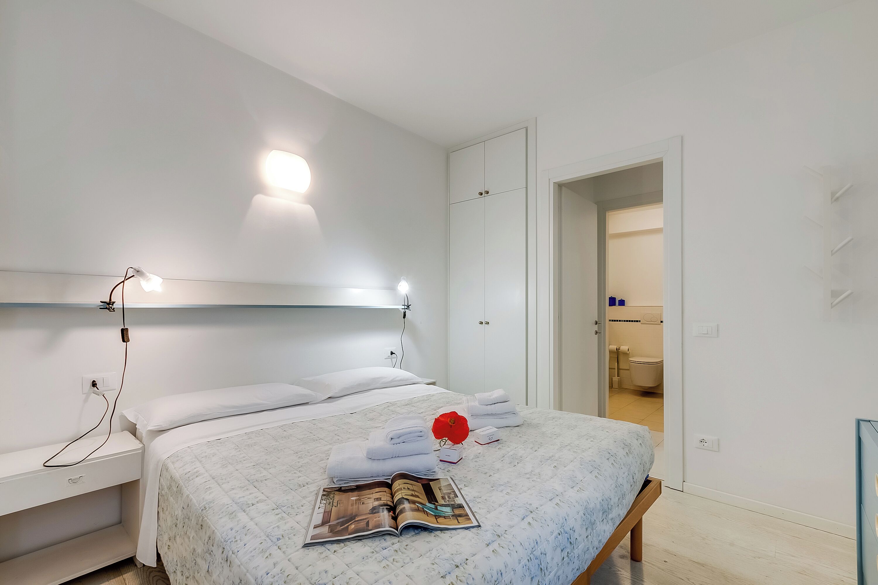 A neatly made bed with white linens and a magazine, a bedside table with a lamp, and a door leading to a bathroom.
