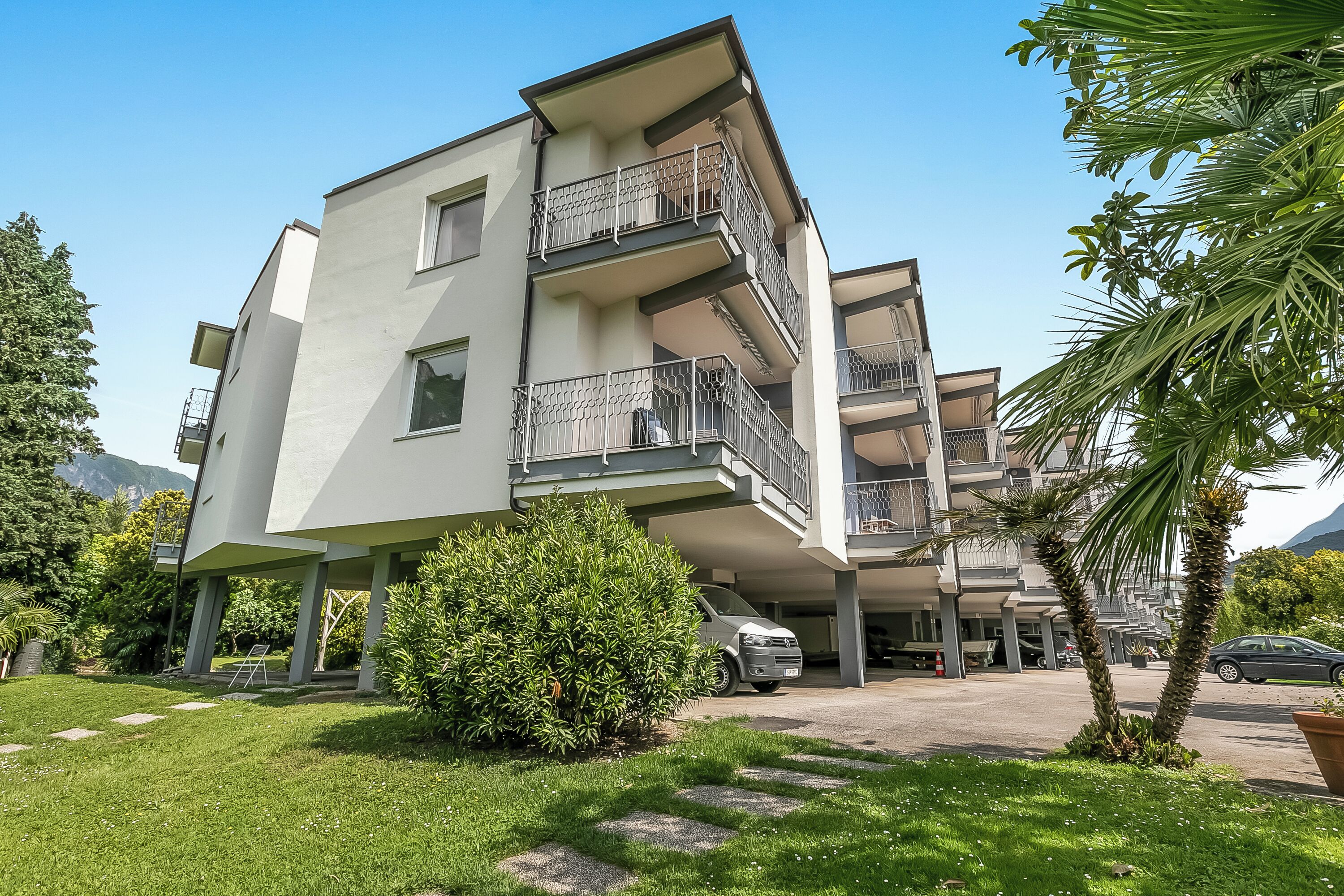 A modern multi-story residential building with balconies, a parking area, and surrounding greenery.
