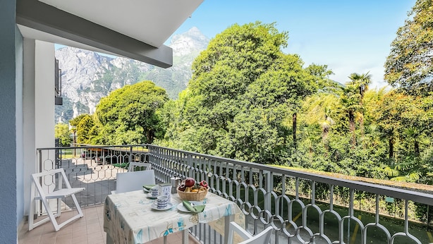 A balcony with a table set for dining, surrounded by a railing, overlooking a lush garden and a mountain. - Riva Lago e Spiaggia Apartment 3 (Riva del Garda)
