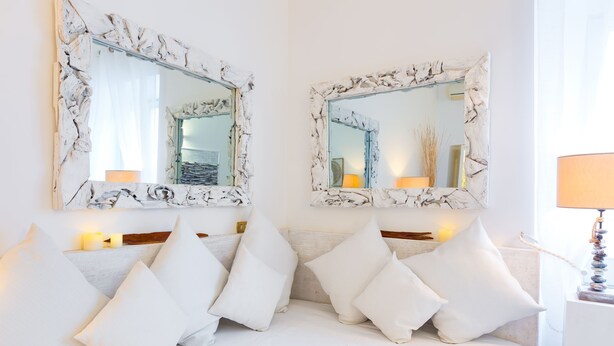 Een wit bed met meerdere kussens en twee grote spiegels met witte lijsten aan de muur.