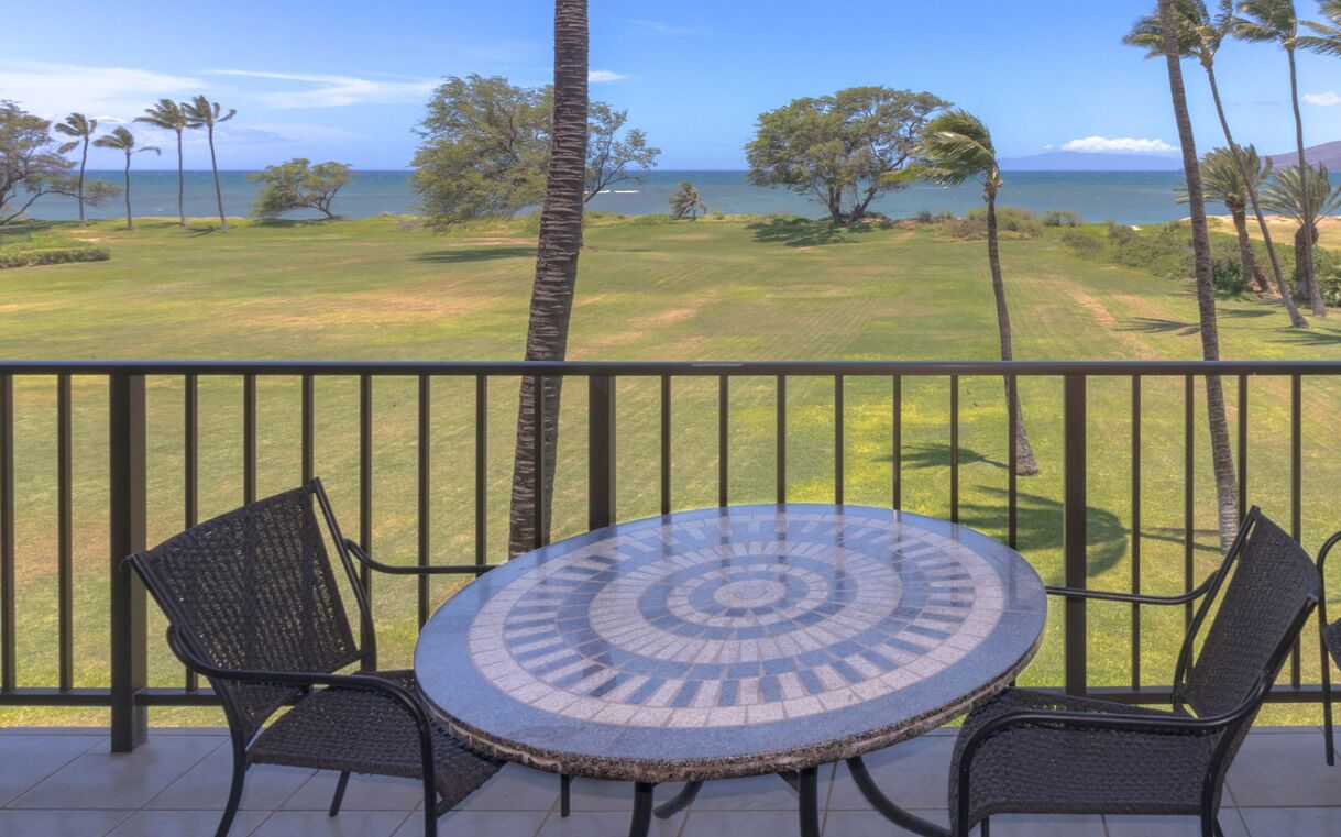 A round table with a mosaic pattern, two wicker chairs, and a view of a golf course and ocean.