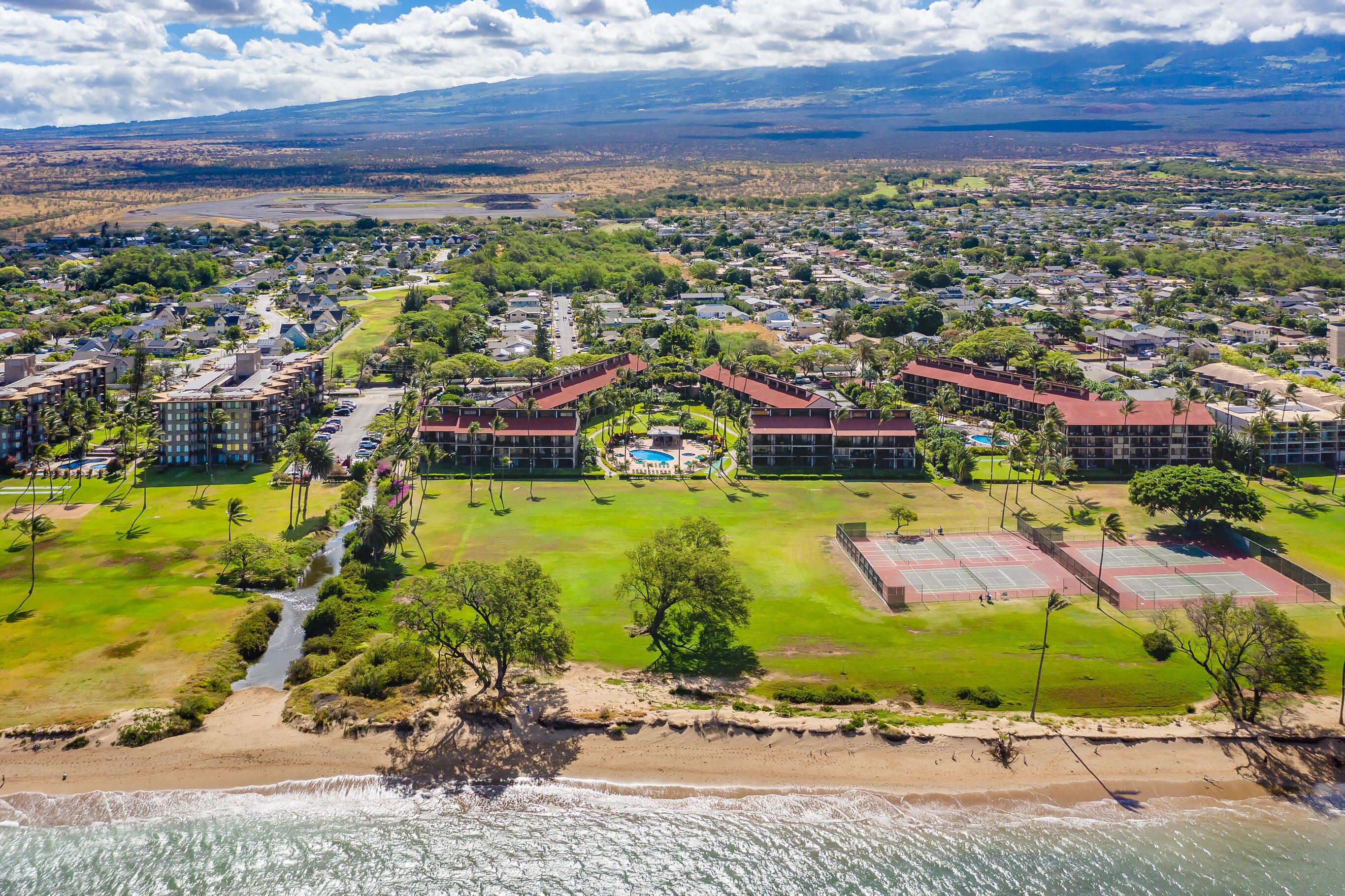 An aerial view of a coastal residential area with a beach, a tennis court, and a swimming pool.