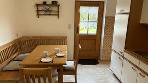 A dining area with a wooden table and bench, a kitchen with white cabinets, and a door leading outside. - Apartment- 2 persons, 80sqm, 1 bedroom, balcony, WLAN (Schleching)