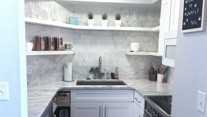 A modern kitchen with white cabinets, a stainless steel sink, and a built-in oven. There are potted plants on the countertop and a welcome sign on the wall.