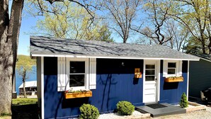 Una casa pequeña con fachada azul oscuro, detalles blancos y techo inclinado. Cuenta con dos ventanas con contraventanas blancas, una puerta principal y un pequeño porche.