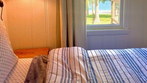 Un dormitorio con una cama, una ventana que da a árboles y un edificio, y una lámpara en la pared.