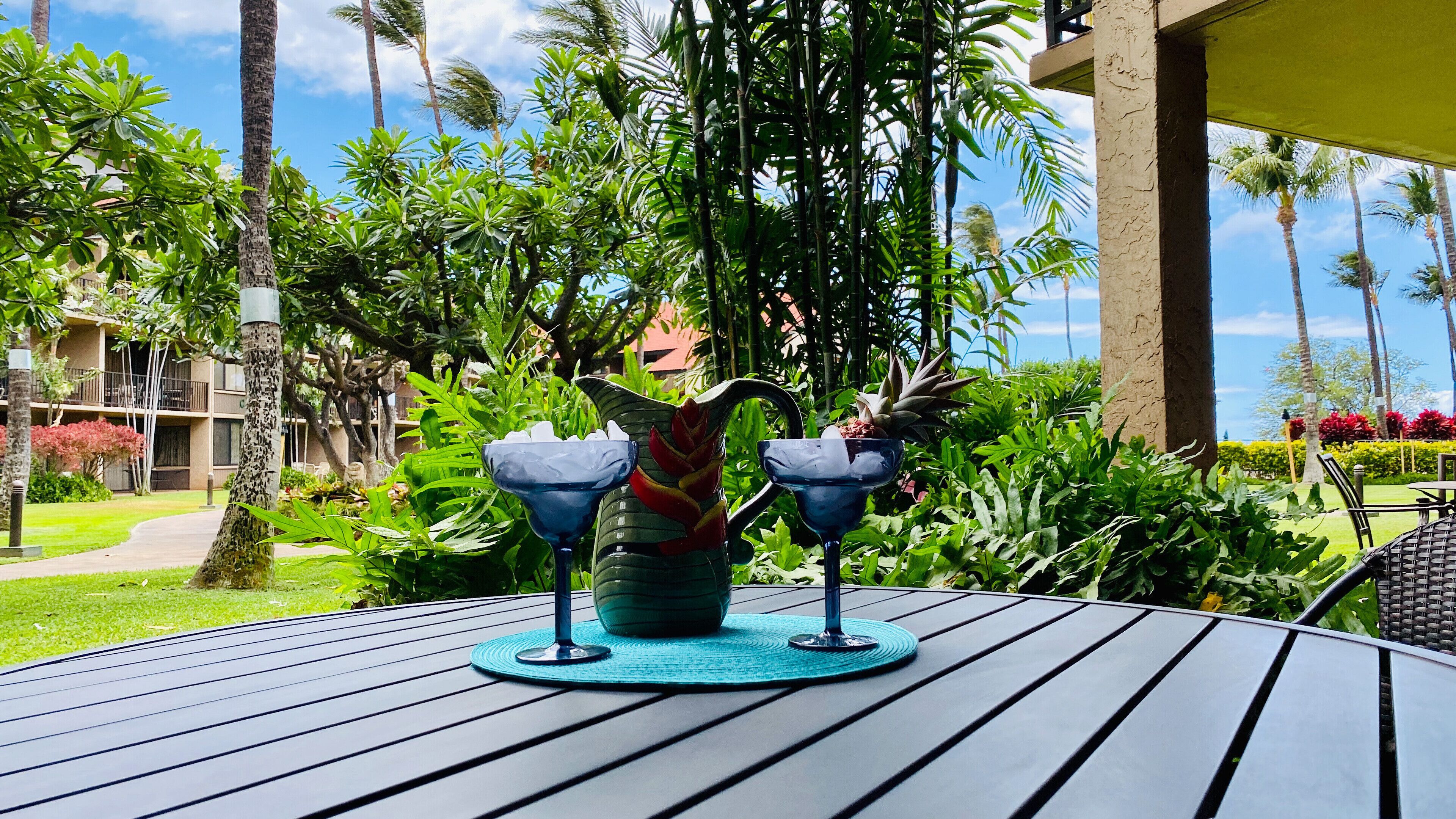 A table with a decorative pitcher and two glass vases, surrounded by lush greenery and palm trees.