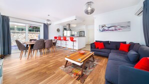 A modern living room with a dark blue sofa, red cushions, and a wooden coffee table. There is a dining area with a wooden table and grey chairs. The room has a large window with curtains, a kitchen in the background, and a colorful rug on the floor.