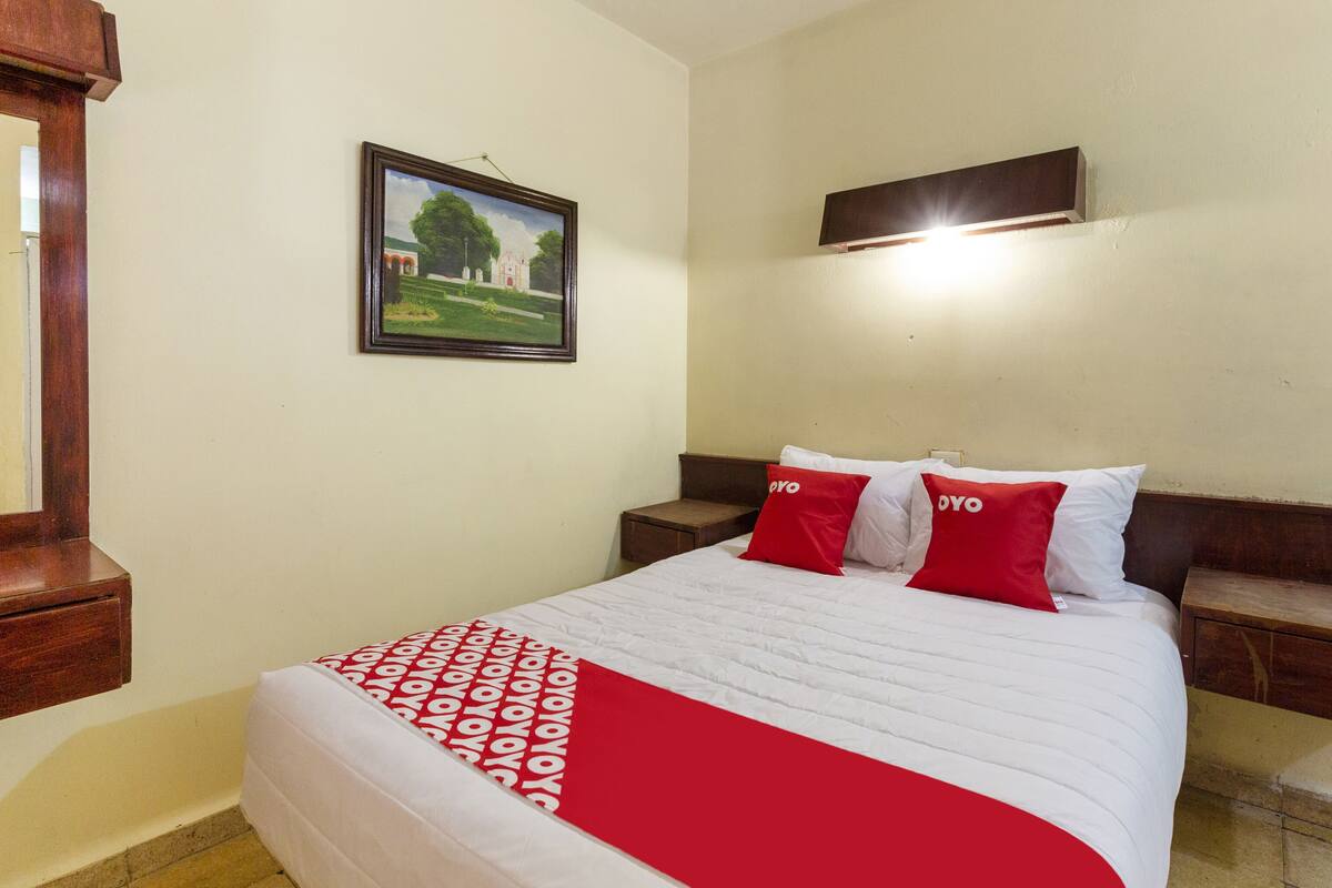 A hotel room with a bed, a nightstand, a wooden headboard, a framed picture on the wall, and a TV mounted above the bed.