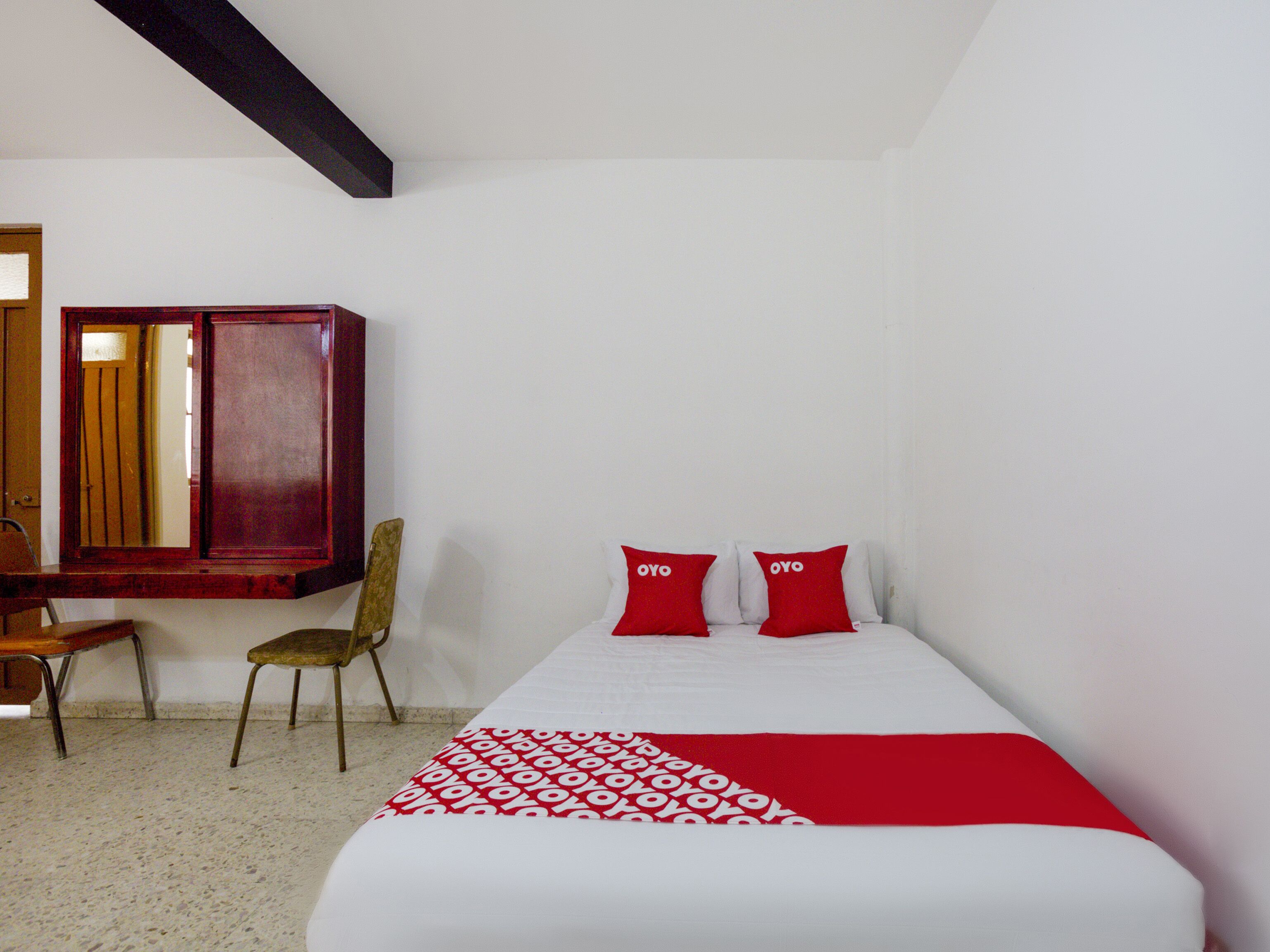 A bedroom with a bed, two red pillows, a wooden chair, a wooden cabinet, and a mirror.