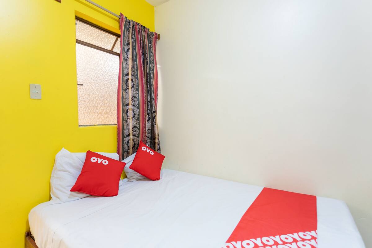 A hotel room with a yellow wall, a bed with white bedding and red pillows, and a window with patterned curtains.