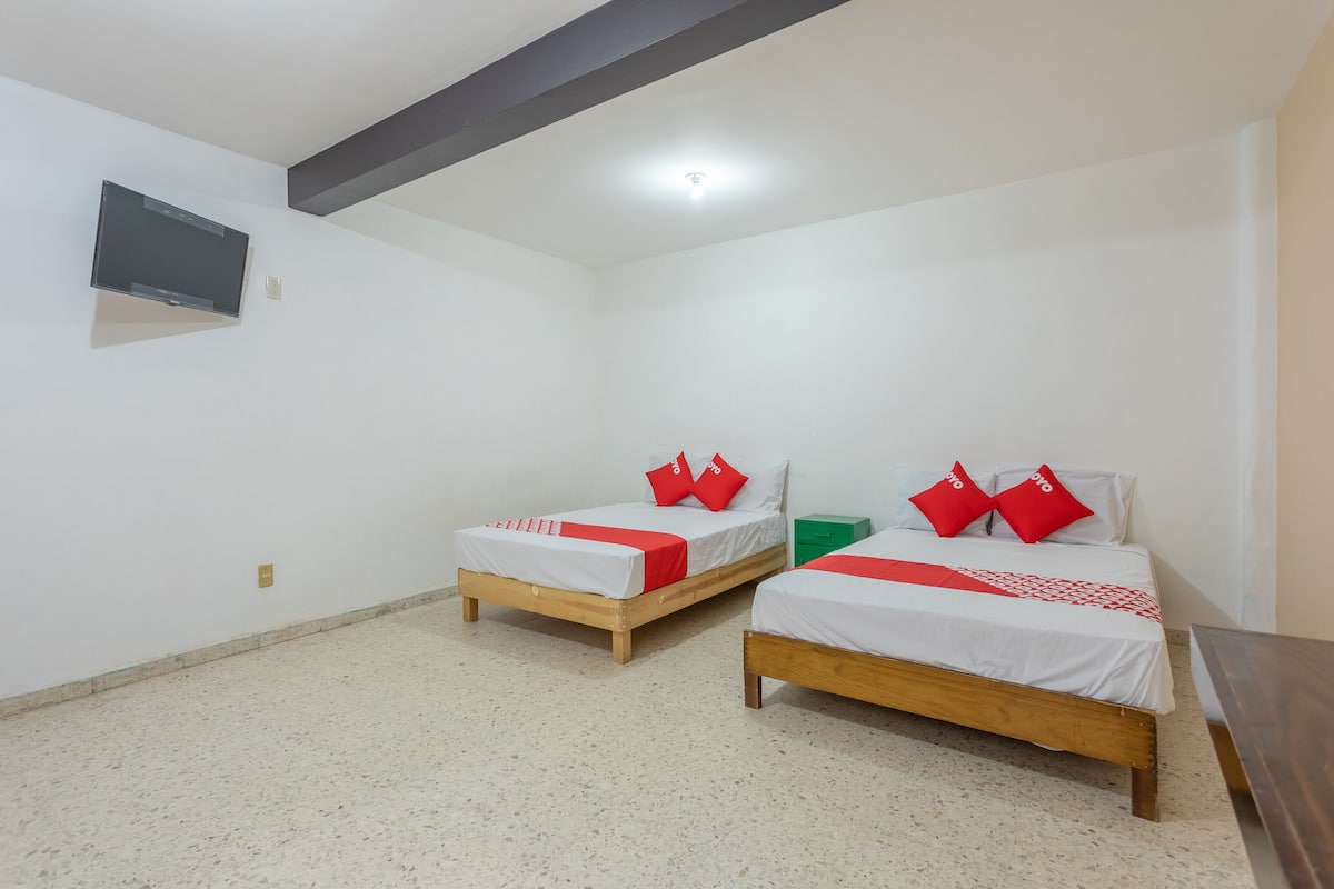 A room with two wooden beds, each with white bedding and red pillows, a green nightstand, a flat-screen TV mounted on the wall, and a wooden bench.