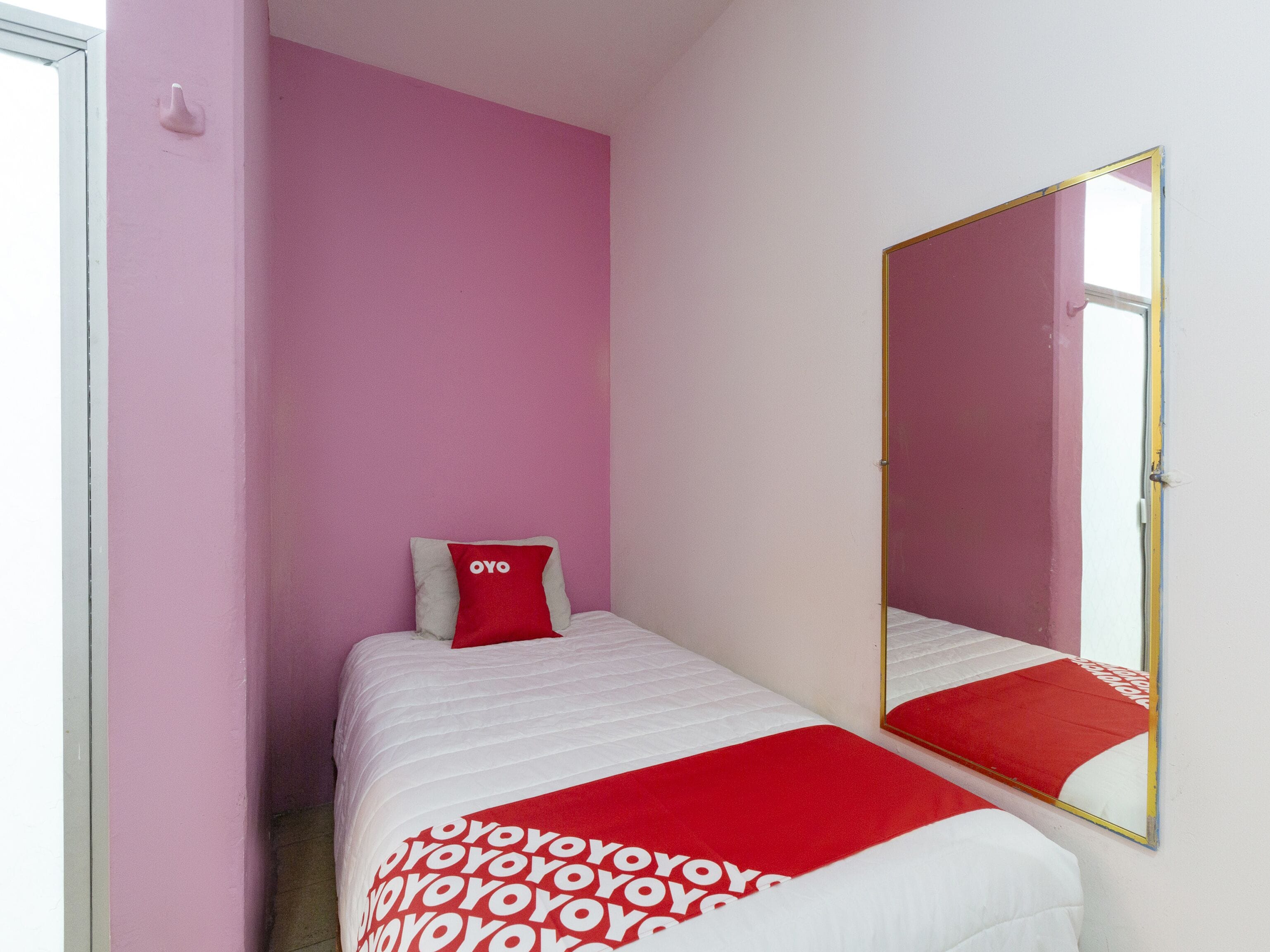 Quarto pequeno com uma cama de solteiro, travesseiro vermelho, espelho e paredes cor-de-rosa.
