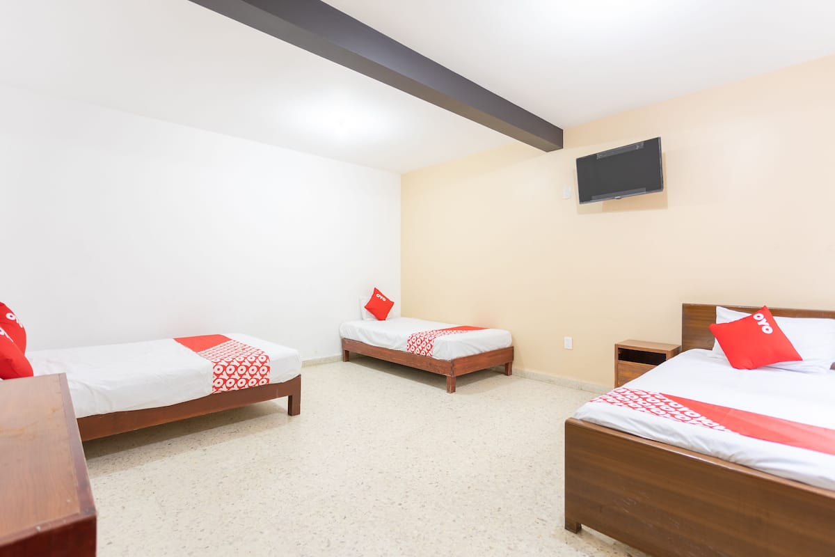 A hotel room with three single beds, each with a red pillow and a white bedspread. There is a wooden nightstand between the beds, and a flat-screen TV mounted on the wall.