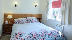 A bedroom with a wooden headboard, a floral bedspread, two wall-mounted lights, a bedside table with a lamp, and a window with a view of flowers.