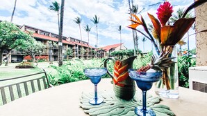 Una mesa con dos vasos azules y un jarrón decorativo con una planta, con el telón de fondo de un patio de hotel con palmeras y un edificio con techo rojo.
