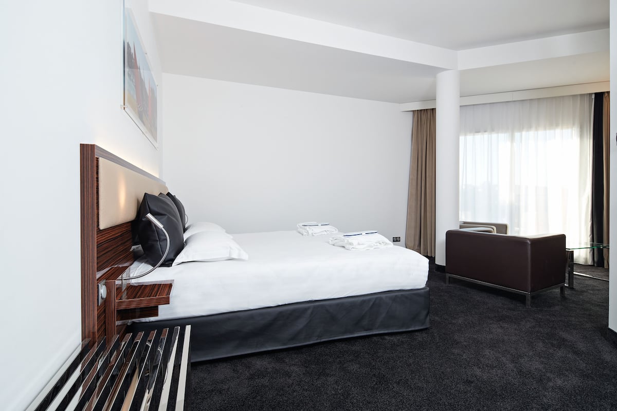 A hotel room with a large bed, a desk, and a chair. There is a window with curtains and a picture on the wall.
