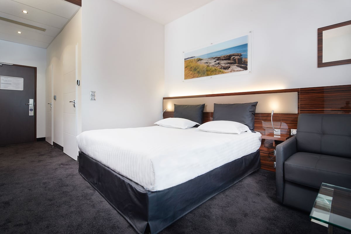 A hotel room with a large bed, a grey armchair, a small table, and a painting on the wall.