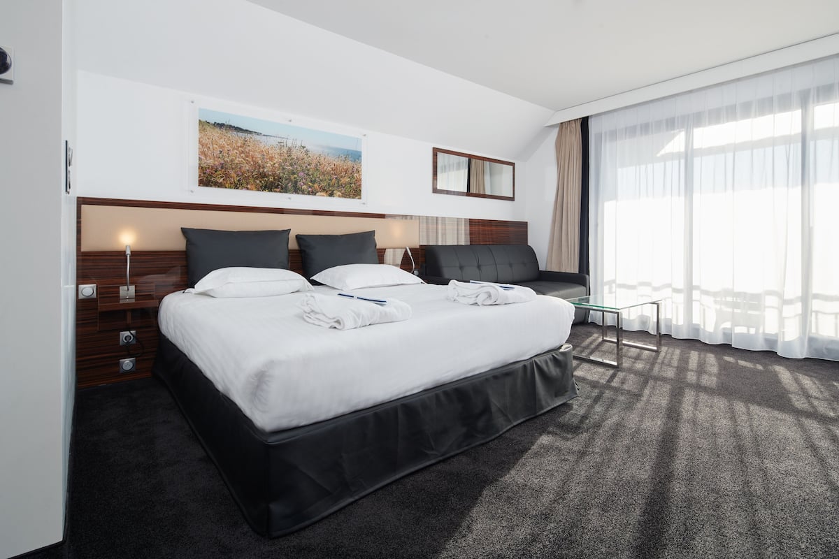 A modern hotel room with a large bed, a bedside table, a lamp, and a view of the outdoors through sheer curtains.