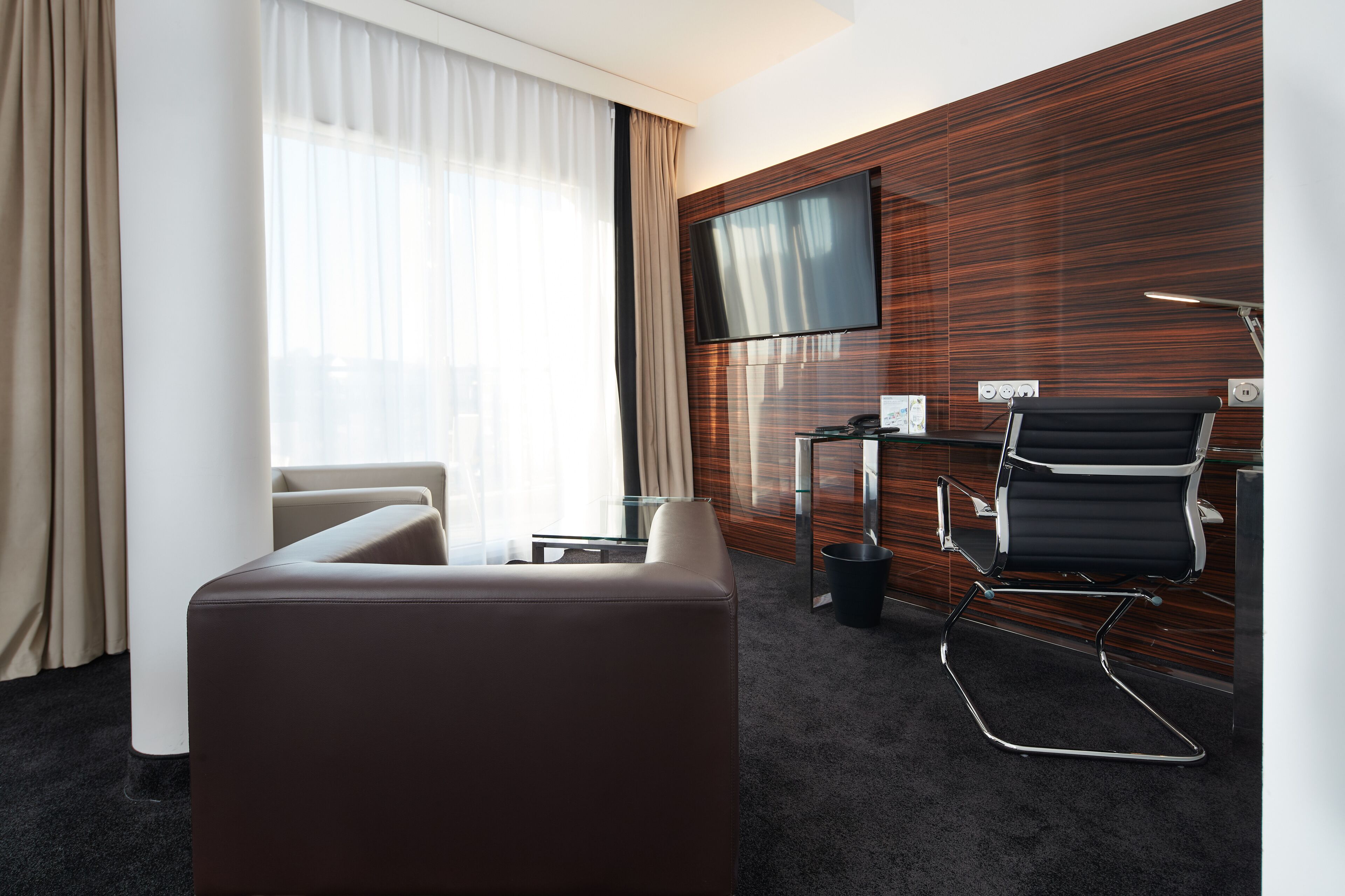 Ein modernes Hotelzimmer mit einer Ledersofa, einem schwarzen Bürostuhl, einem Flachbildfernseher, der an einer Holzwand montiert ist, und einem kleinen Tisch mit einer Lampe.