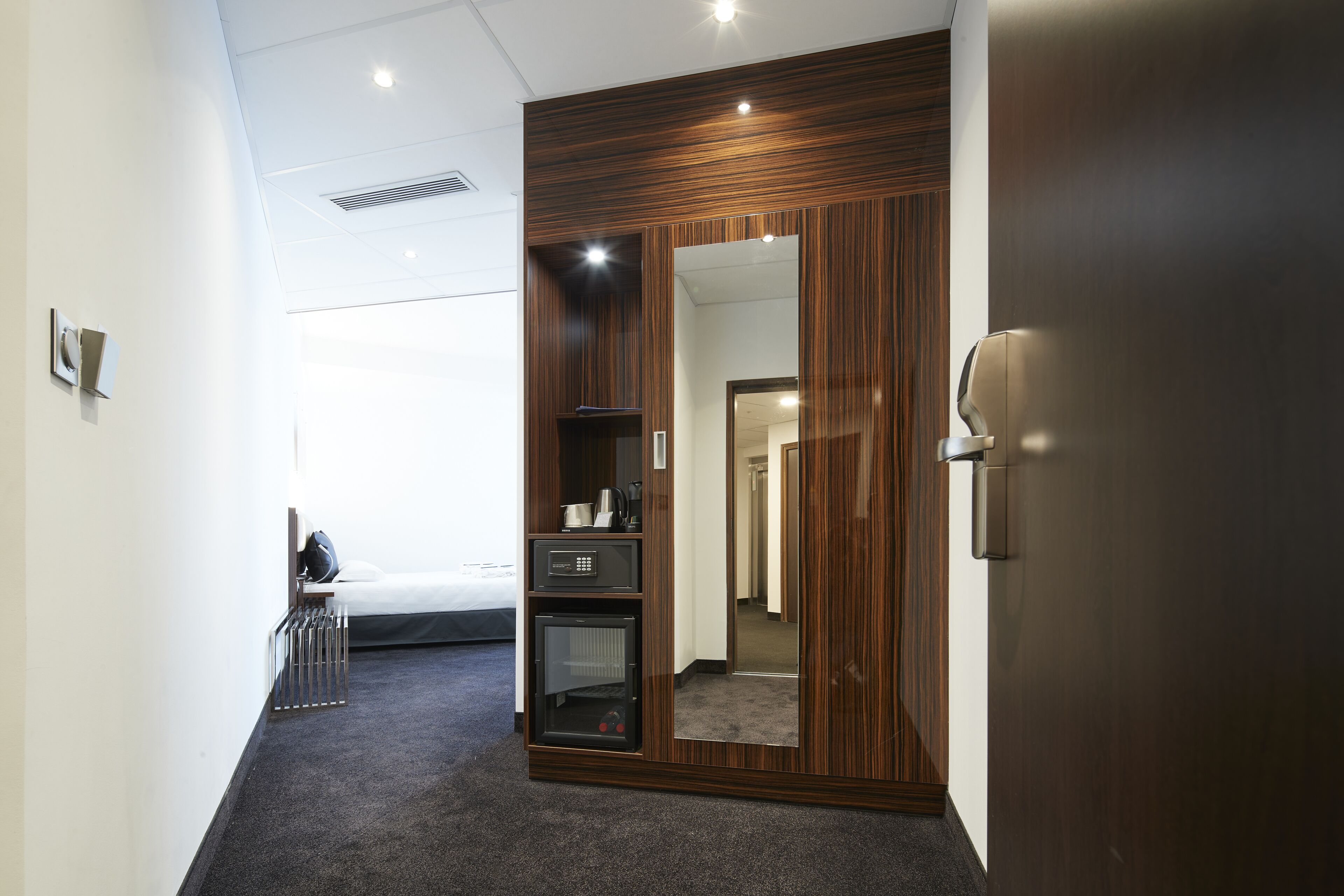 A hotel room with a wooden wardrobe, a bed, a small kitchenette, and a mirror.