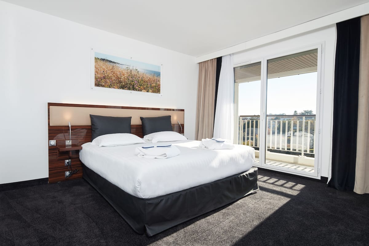 A hotel room with a large bed, a wooden headboard, a nightstand, a window with a view of a building, and a balcony with a railing.