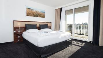 Habitación de hotel con una cama grande, cabecera de madera, mesita de noche, ventana con vista a un edificio y balcón con barandilla.