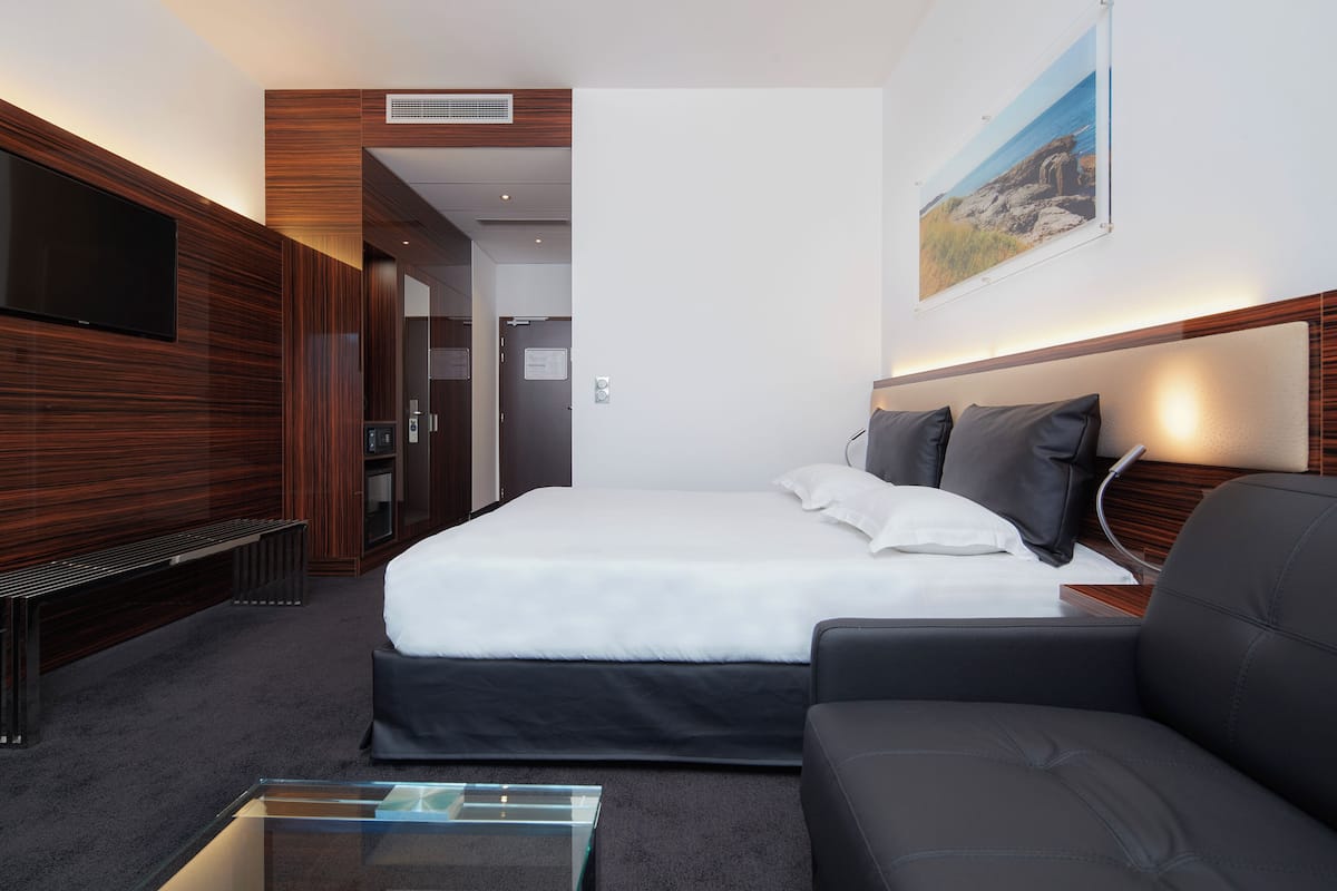 A modern hotel room with a large bed, a sofa, a flat-screen TV, and a glass coffee table.