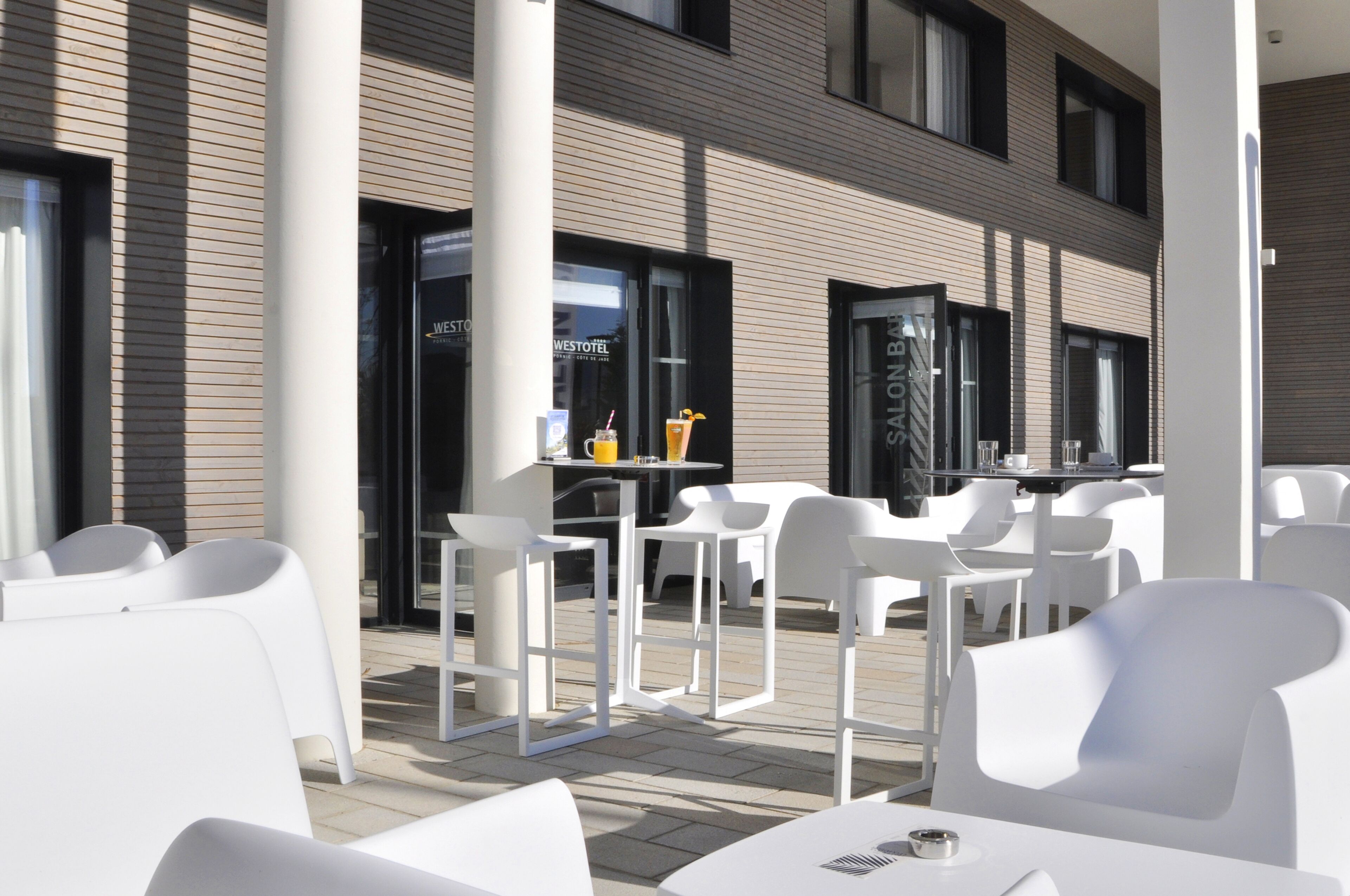An outdoor seating area with white chairs and tables, a bar area with a drink, and a building with a modern design and large windows.