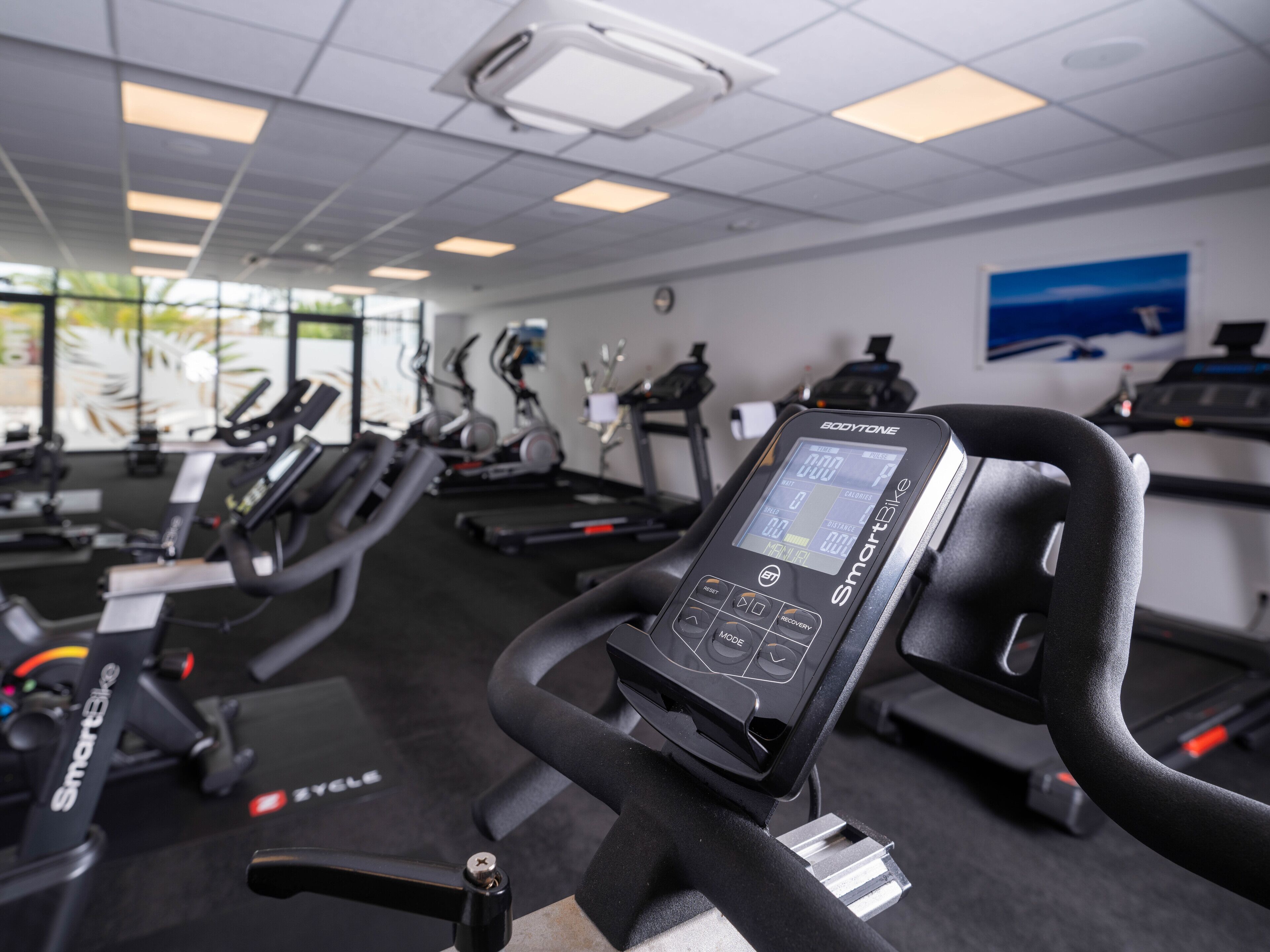 Un gimnasio con varias máquinas de ejercicio, incluyendo una cinta de correr y bicicletas estáticas, y una pantalla digital en una de las máquinas.