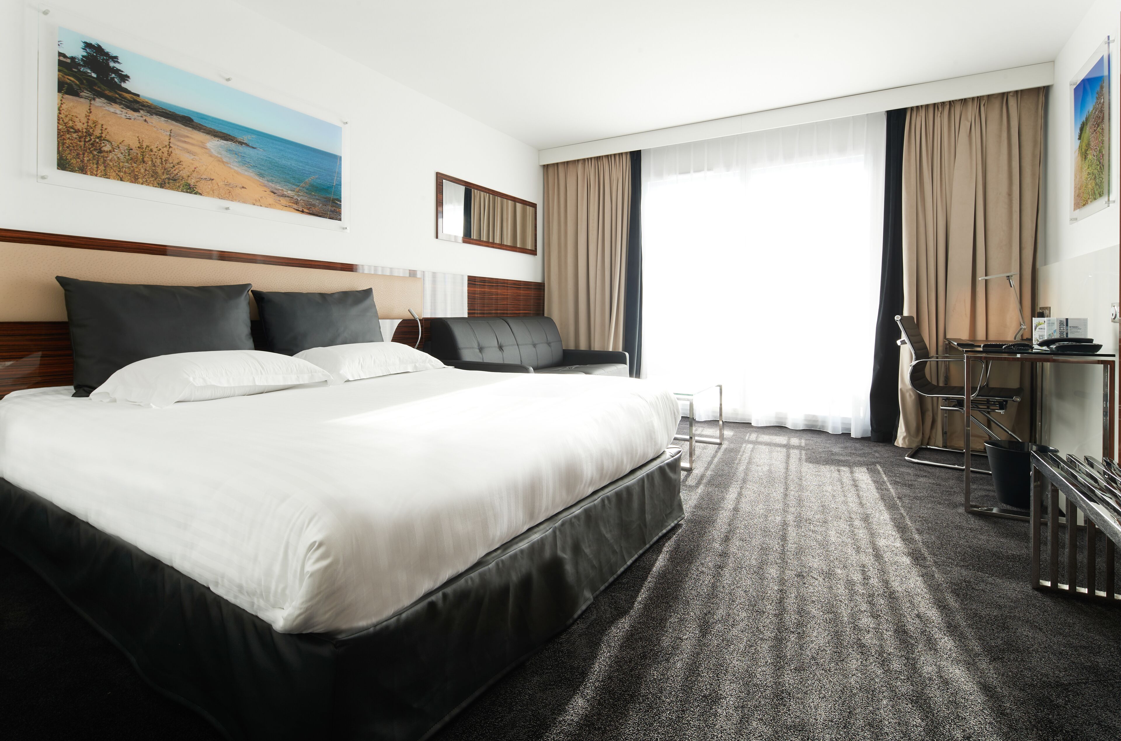 Habitación de hotel con una cama grande, un escritorio con silla, una computadora portátil y un ventanal con cortinas.