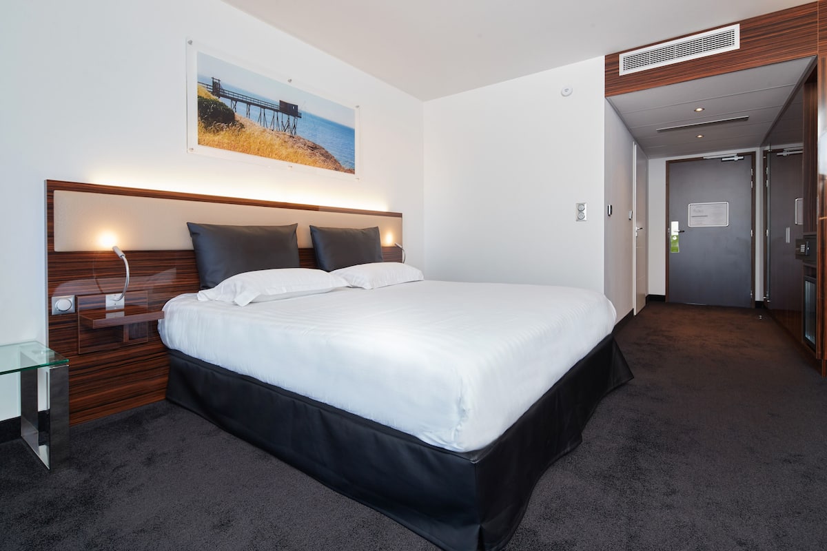 A modern hotel room with a large bed, a bedside table, a wall-mounted picture, and a door leading to another room.