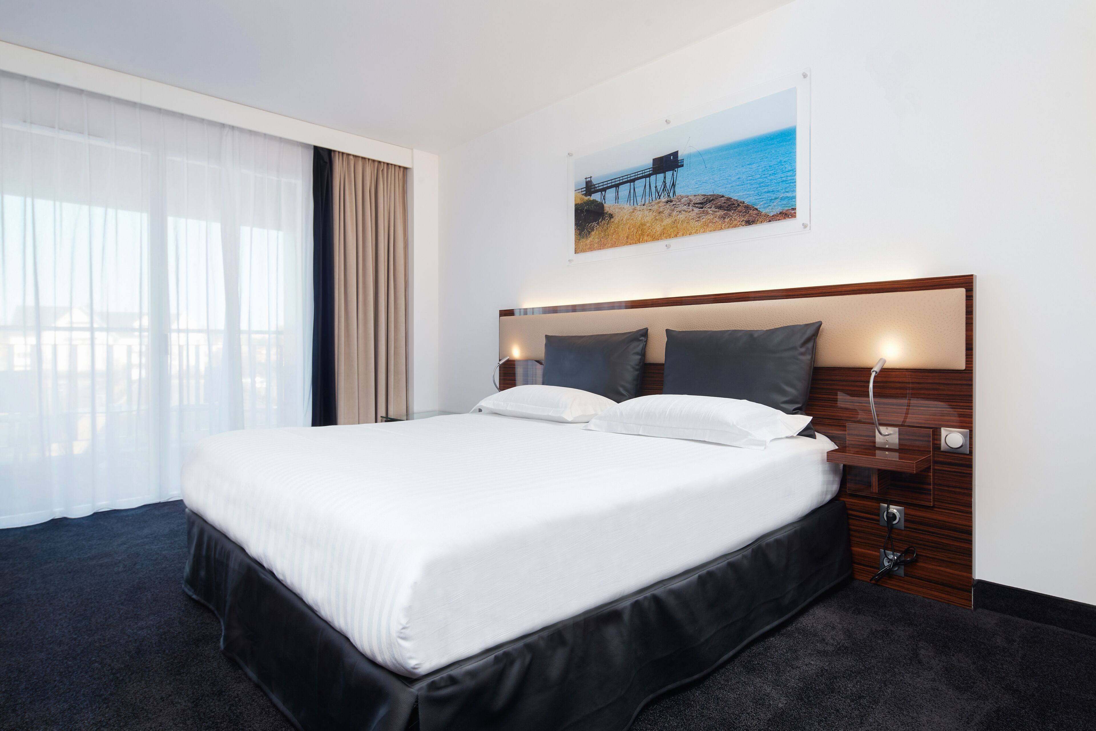 A hotel room with a large bed, a wooden headboard, a nightstand, and a window with sheer curtains.