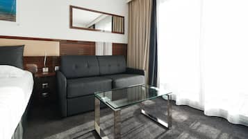 A modern hotel room with a bed, a sofa, a glass coffee table, a nightstand, a lamp, a mirror, and a window with curtains.