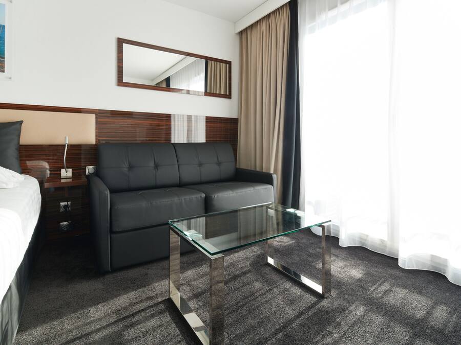 A modern hotel room with a bed, a sofa, a glass coffee table, a nightstand, a lamp, a mirror, and a window with curtains.