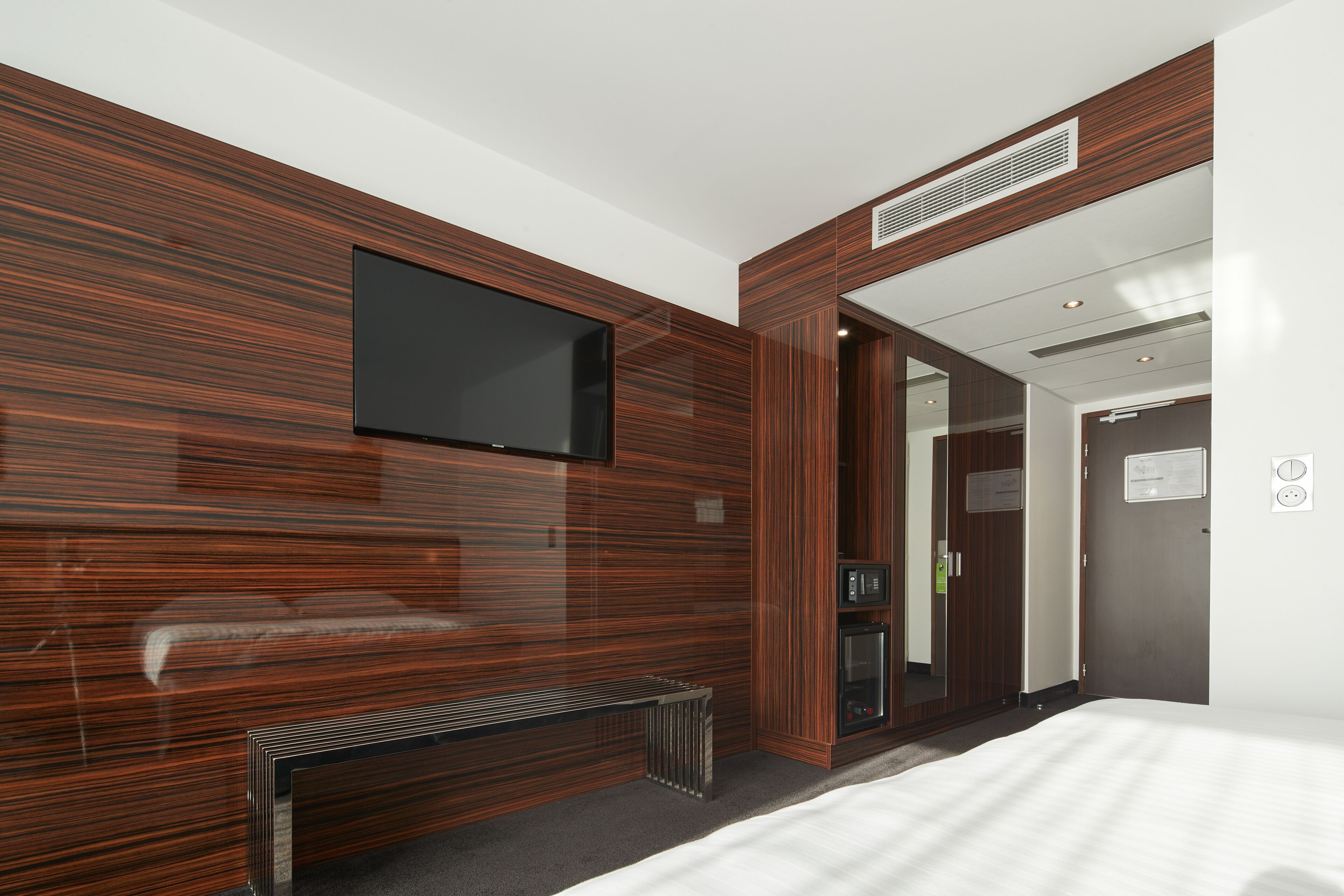 A modern hotel room with a large flat-screen TV mounted on a wooden wall, a bench, and a door leading to a bathroom.