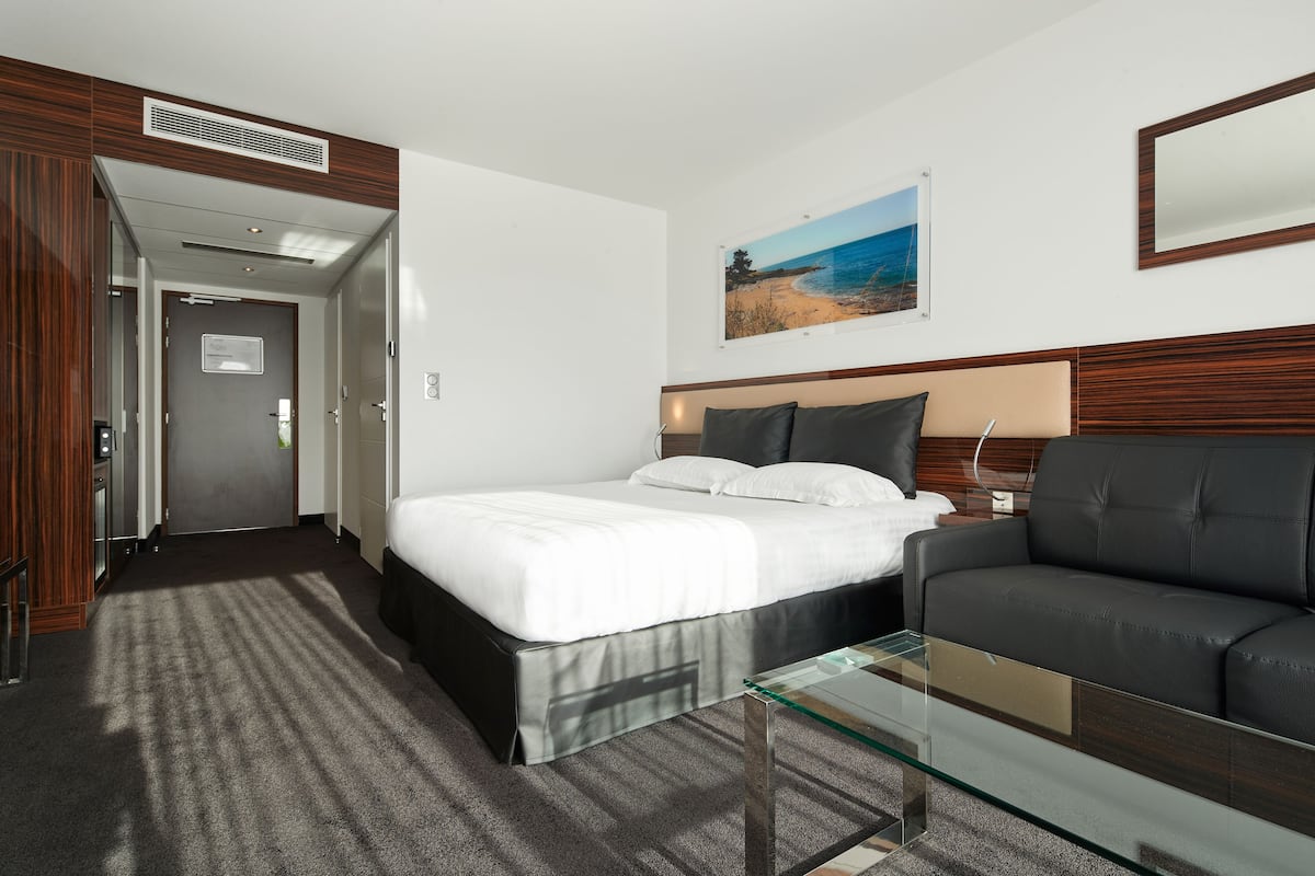 A hotel room with a bed, a sofa, a glass coffee table, a painting of a beach, and a wooden headboard.