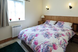 A bedroom with a wooden headboard, a floral bedspread, a bedside table with a lamp, and a window with curtains.