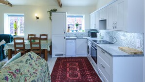A compact kitchen with white cabinets, a washing machine, and a microwave. There is a dining area with a table and chairs, and a red rug on the floor. -  YORK single storey cottage ,gardens & open views.Thicket Priory & Derwent Ings. (Thorganby, York)