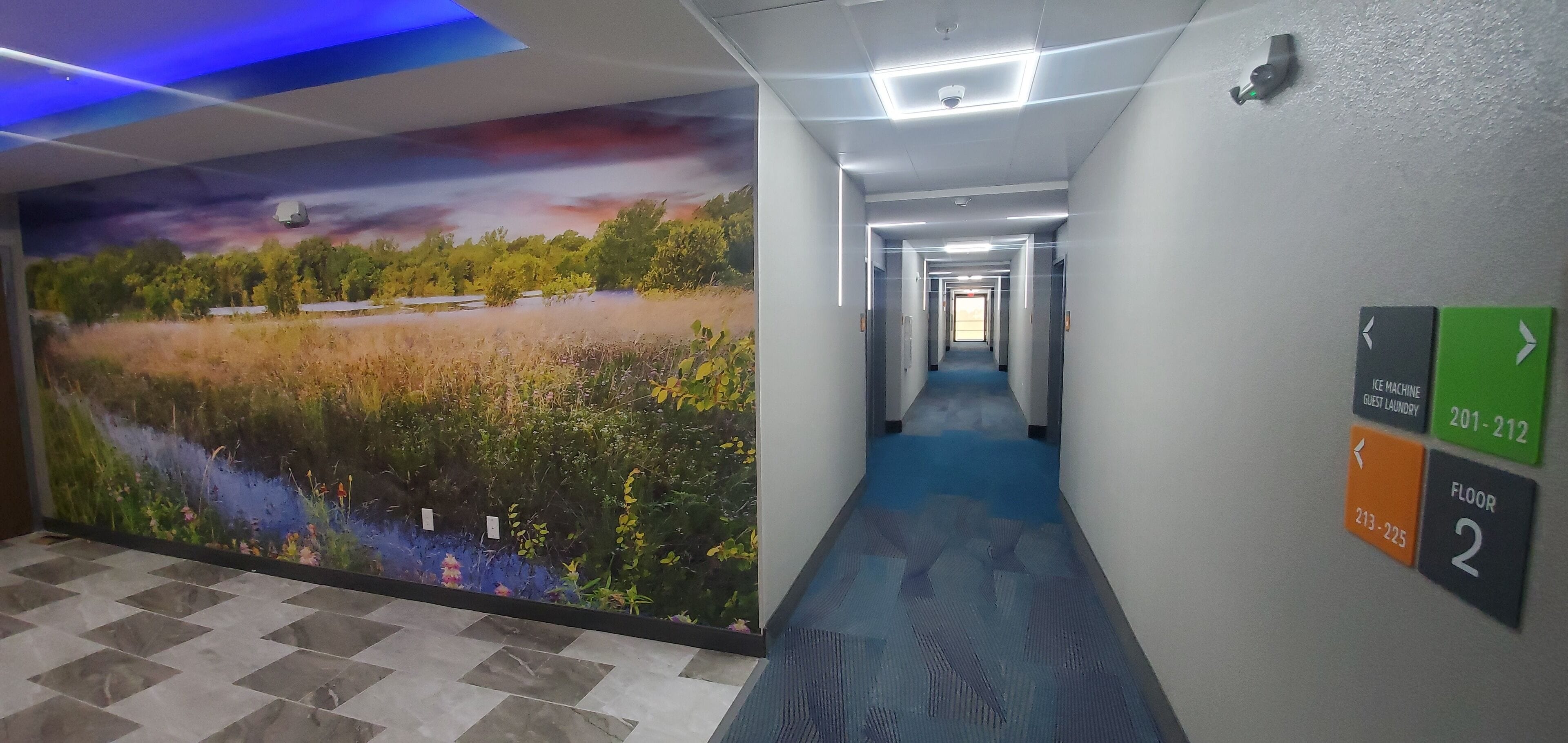 A hotel corridor with a mural of a natural landscape, directional signs for the ice machine and guest laundry, and a blue carpet.