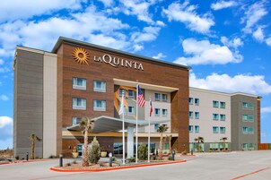 A La Quinta hotel with a modern design, featuring a prominent sun logo, multiple windows, and a well-maintained entrance area with palm trees and a flag.