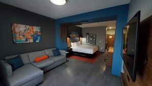 Une chambre d’hôtel moderne avec un canapé gris, un lit, un téléviseur à écran plat et un tableau au mur.