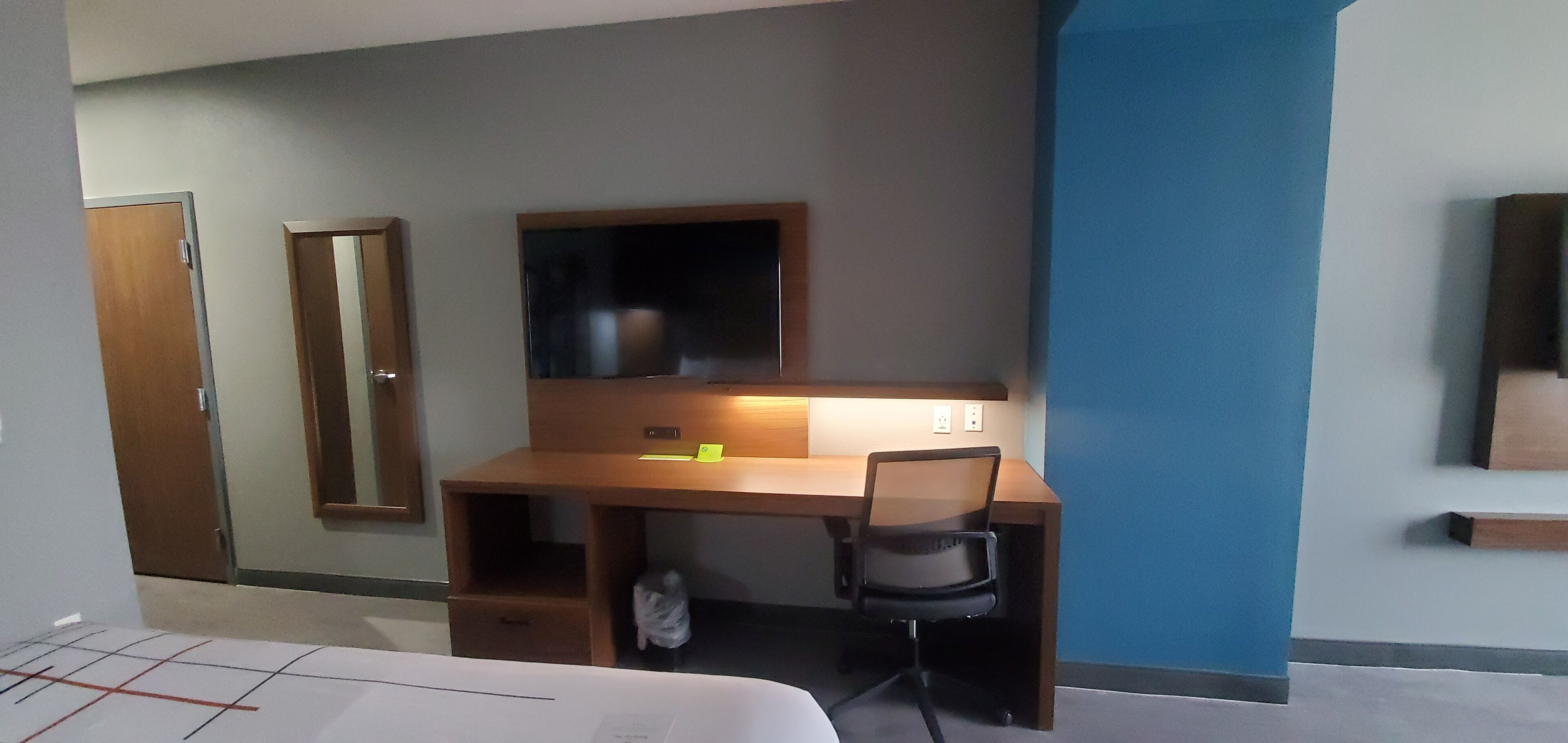 A hotel room with a wooden desk, a chair, a bed, a wardrobe, and a mirror.