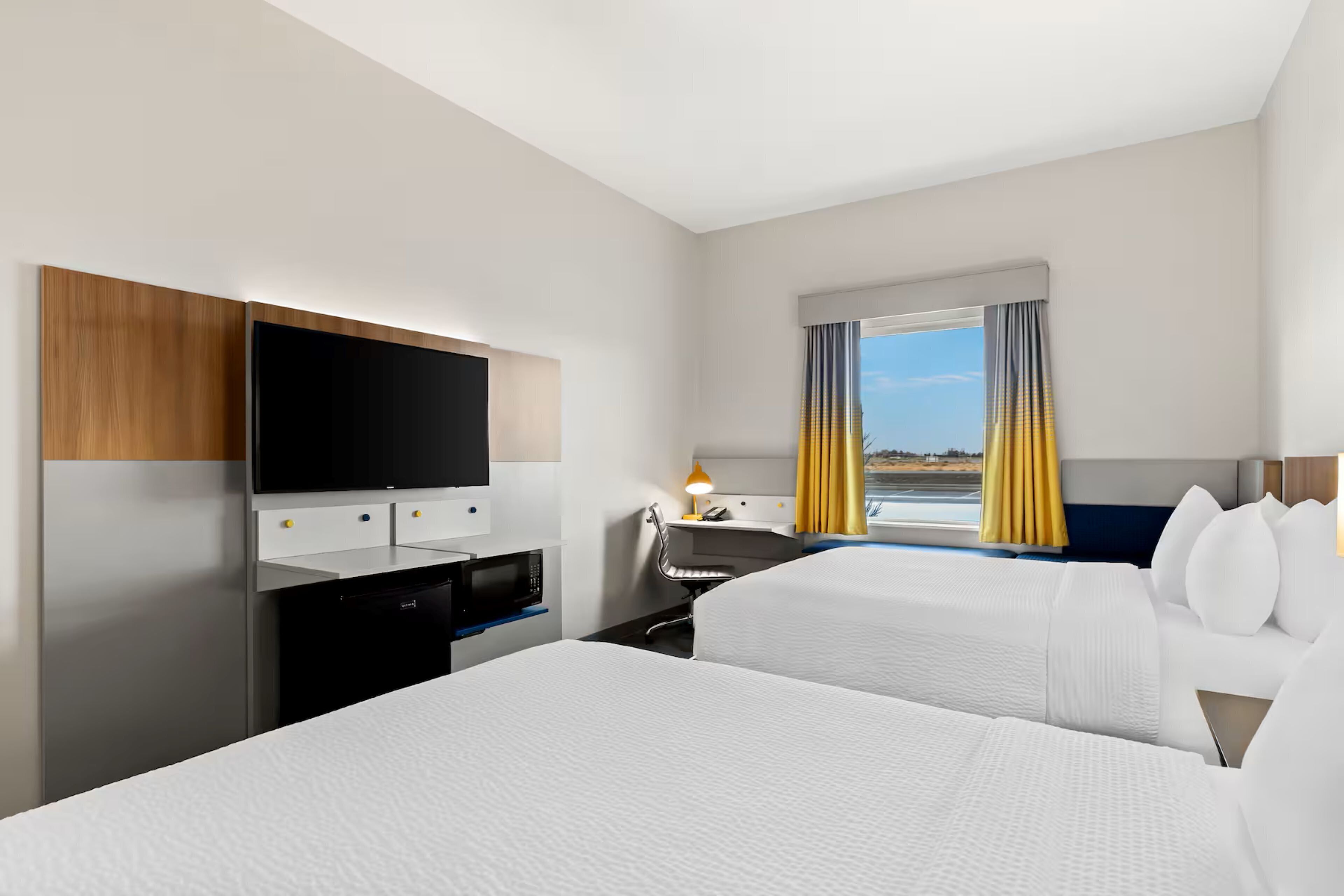 A hotel room with two beds, a flat-screen TV, a desk with a lamp, and a view of the outdoors.
