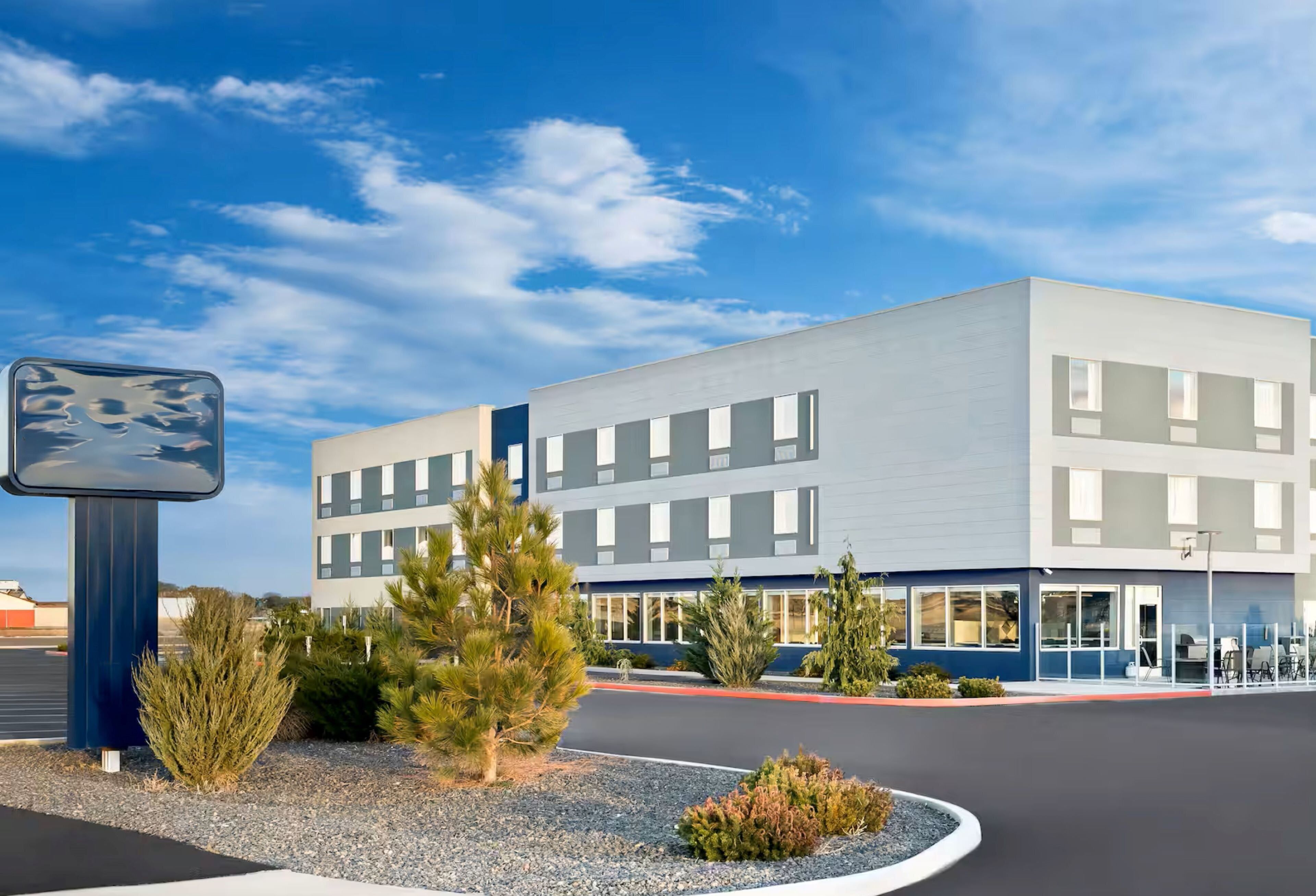 A modern building with a flat roof, rectangular windows, and a parking lot with a few trees and shrubs.